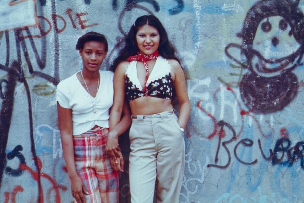 Two teenage girls posing in front of a wall of graffiti in Lynch Park, Brooklyn, 1974.