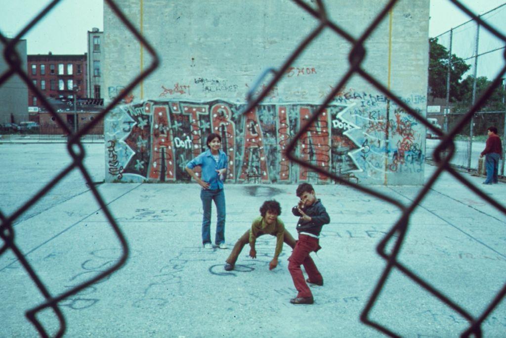 Three boys playing from behind a chain link fence in Brooklyn's Lynch Park, graffiti "A Train" in the background, 1974.
