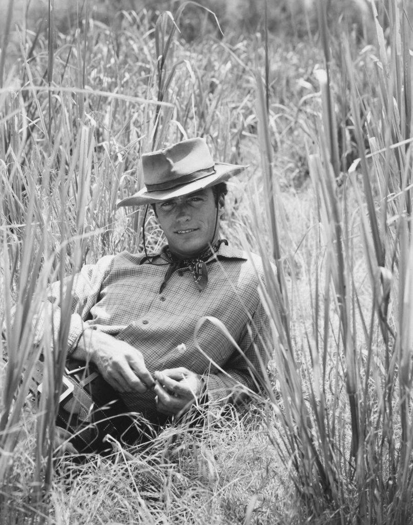 Clint Eastwood smiles as he lies in tall grass for a portrait wearing a cowboy hat and Western outfit, 1961.