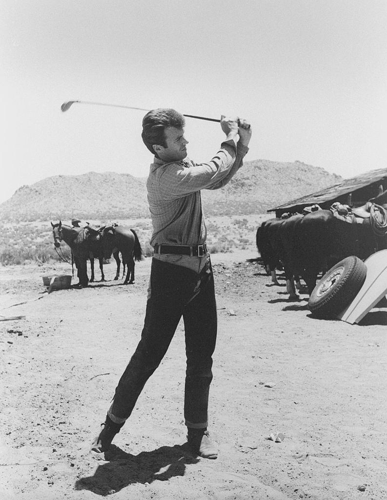 Clint Eastwood wears chaps while he practices his golf swing as a horse looks on during a break in the shooting, 1962.