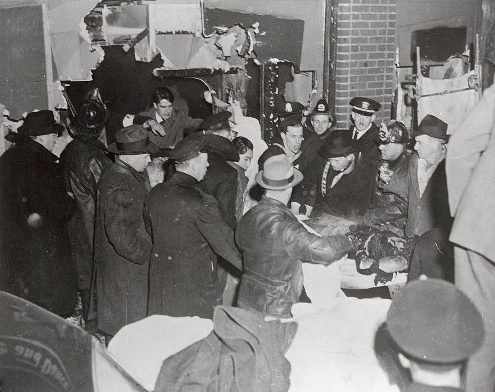 A group of firemen, civilians and uniformed men stand around victim of tragic fire as priest administers last rites.