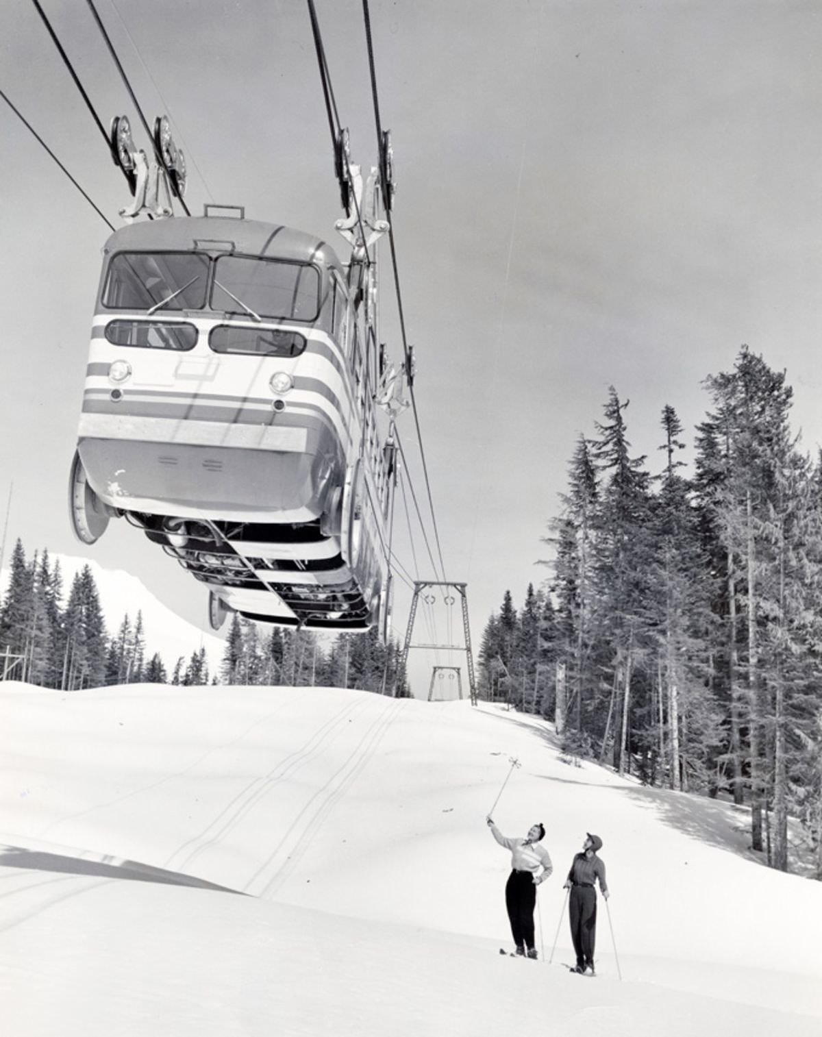 Two skiers stand on a snowy ski slope below a Skiway tram car on November 25, 1951.