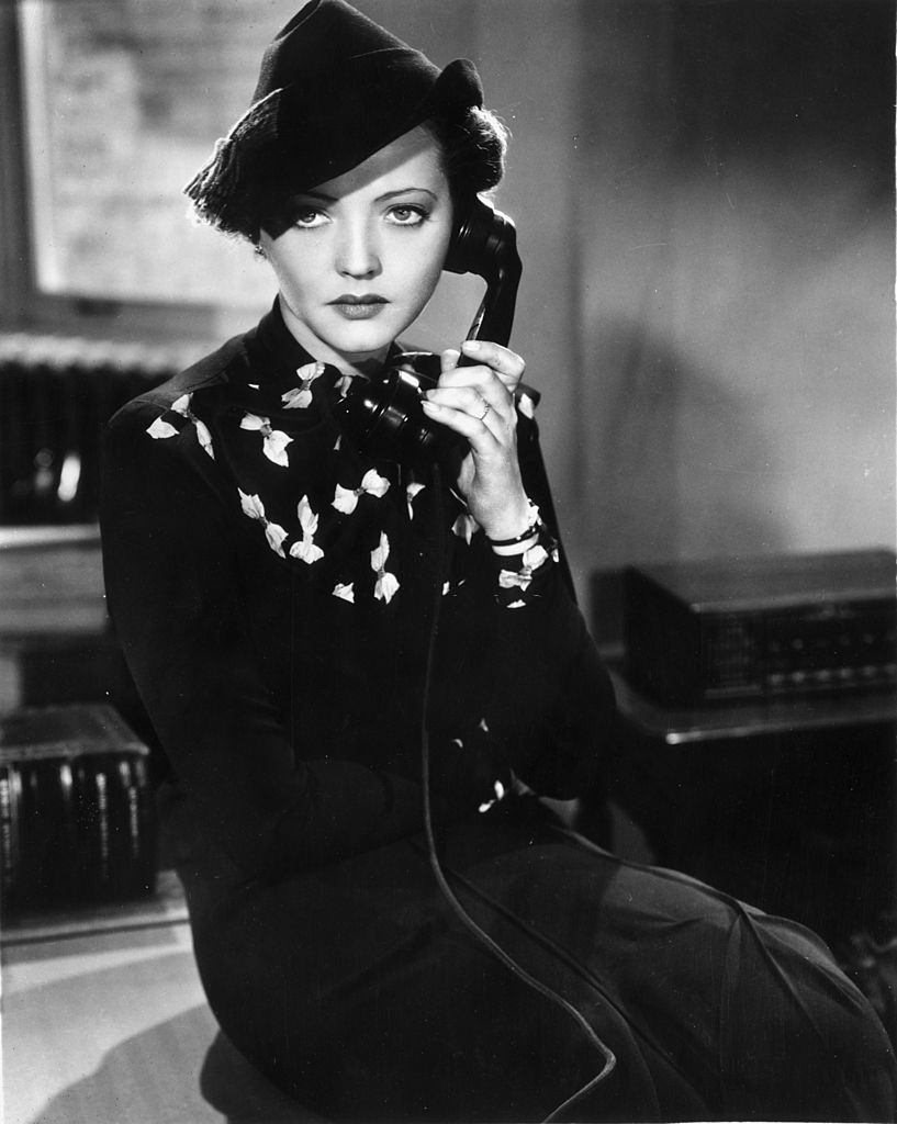 Sylvia Sidney holds a telephone receiver to her ear while sitting on the edge of a desk, 1937.