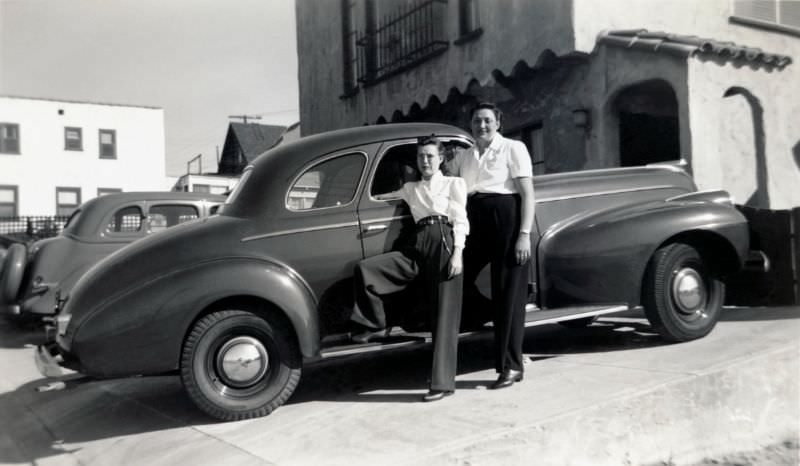 Two ladies posing with a 1940 Oldsmobile Club Coupe in front of a house in Spanish Colonial style in bright sunlight, 1940