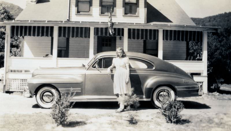 A young lady in a white summer dress posing with a 1941 Oldsmobile Dynamic Cruiser Club Sedan in front of a white timber house, 1941