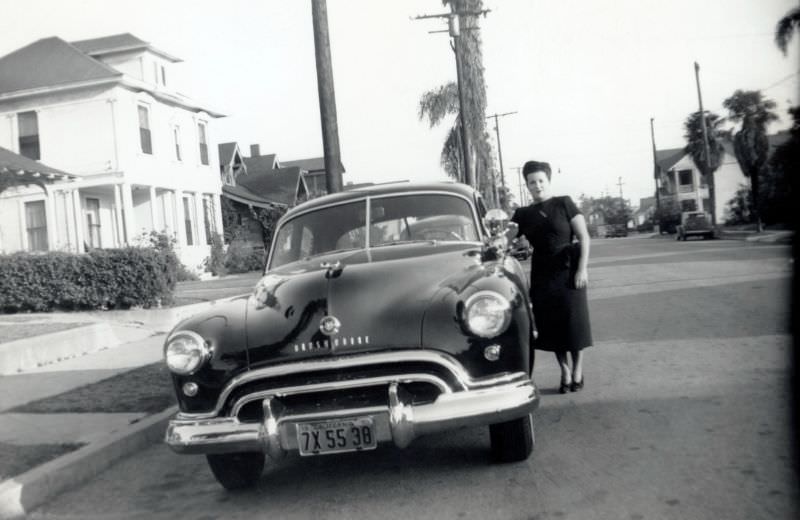 A brunette lady posing with a 1948 Oldsmobile in a suburban street.