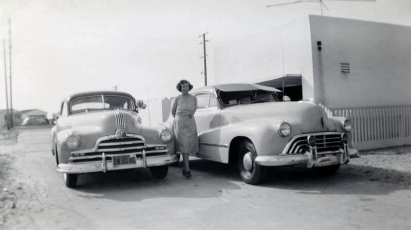A middle-aged lady posing with a pair of 1947 Pontiac and 1947 Oldsmobile in a dusty street on the outskirts of town.