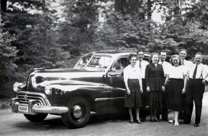 Seven members of a Belgian middle-class family posing with a 1947 Oldsmobile in the countryside, 1948