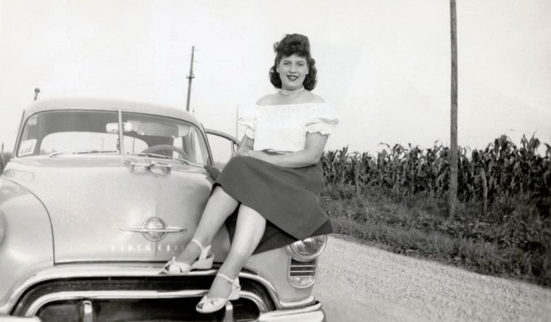 A brunette lady posing on the fender of a 1949 Oldsmobile 88 on a gravel road in the cornfields, somewhere in rural America, 1950