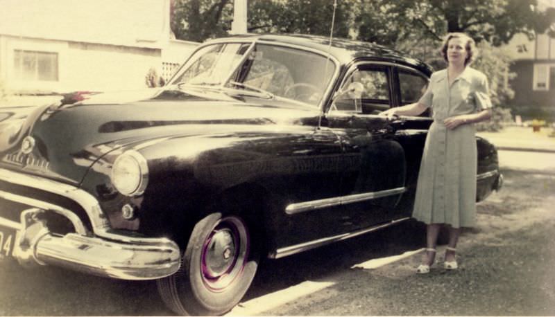 A brunette lady wearing a green 3/4-length dress and white peep-toe sandals posing with a 1948 Oldsmobile in the drive of a suburban home.