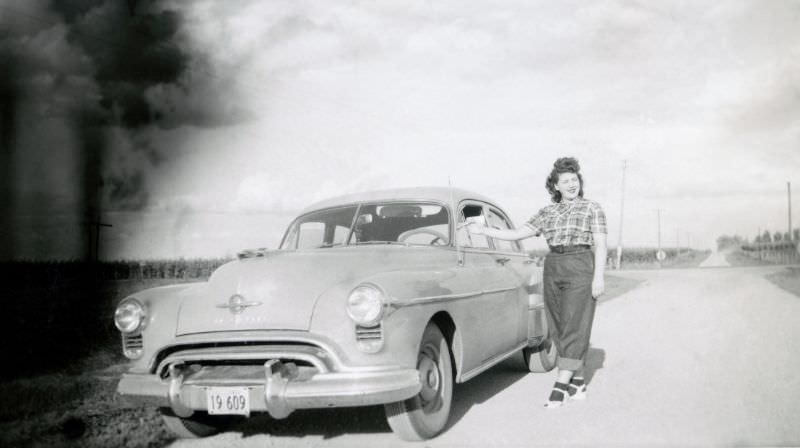 A cheerful lady posing with a 1950 Oldsmobile 76 on a dirt road in the countryside.