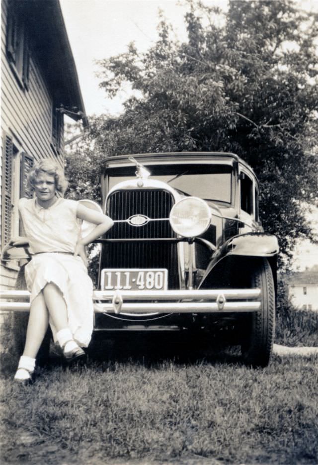 A blonde lady in a white dress posing on the bumper of a 1930 Oldsmobile Six, adorned with an aftermarket propeller hood ornament,1930