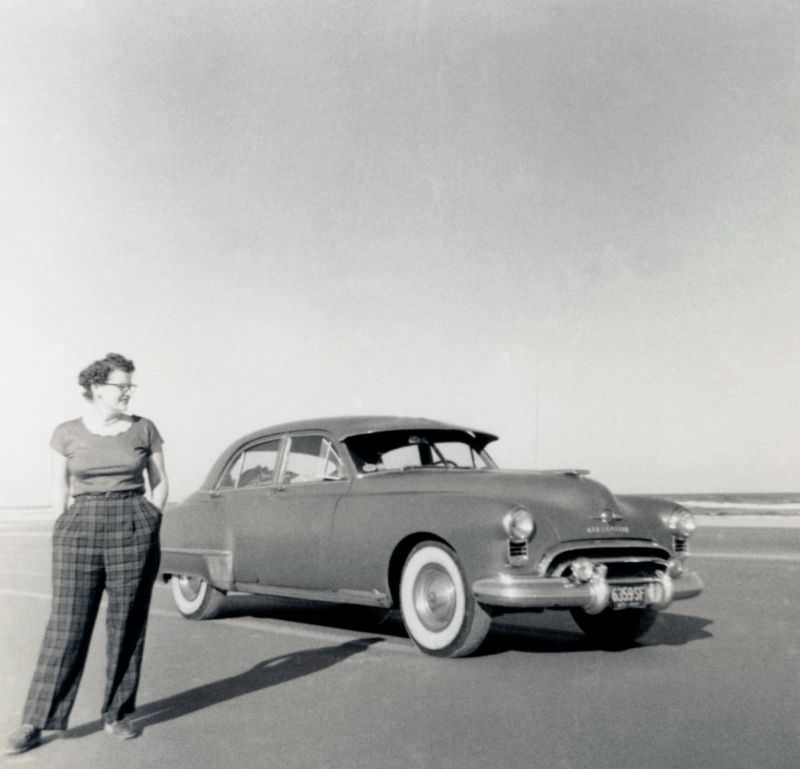 A curly-haired lady dressed in tartan check trousers posing with a 1949 Oldsmobile 88 4-Door Sedan in late afternoon sunshine, 1952