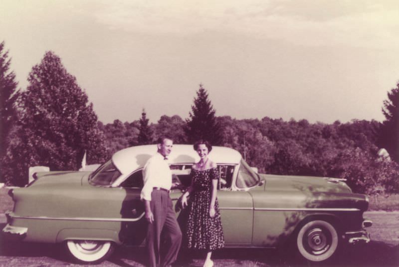 A fellow in a white shirt and a lady in a floral dress posing with a green and white 1954 Oldsmobile Super 88 Holiday Coupe in the countryside.
