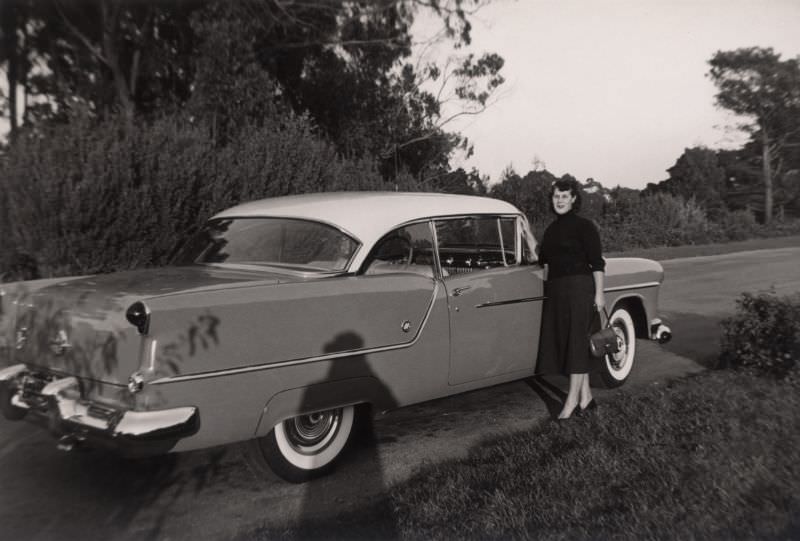 A lady with a handbag posing next to a 1954 Oldsmobile Super 88 Holiday Coupe.