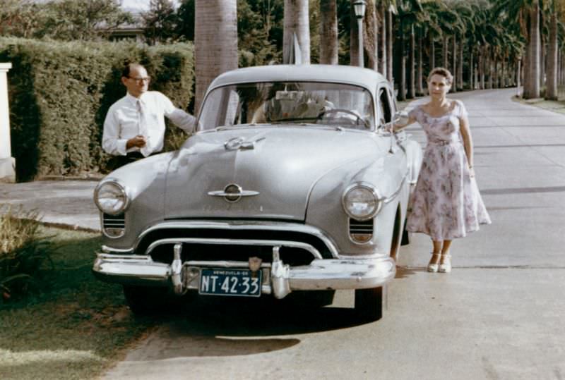A stylish lady in a summer dress and a fellow in a white shirt posing with a 1950 Oldsmobile 88 on an avenue lined with palm trees.
