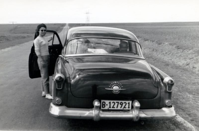 A stylish young lady posing with a 1952 Oldsmobile 98 on the side of a long, straight road running through a plain landscape