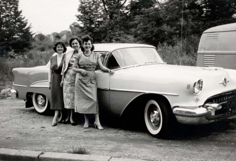 Three cheerful ladies posing with a shiny two-tone 1955 Oldsmobile 88 in the countryside, 1955