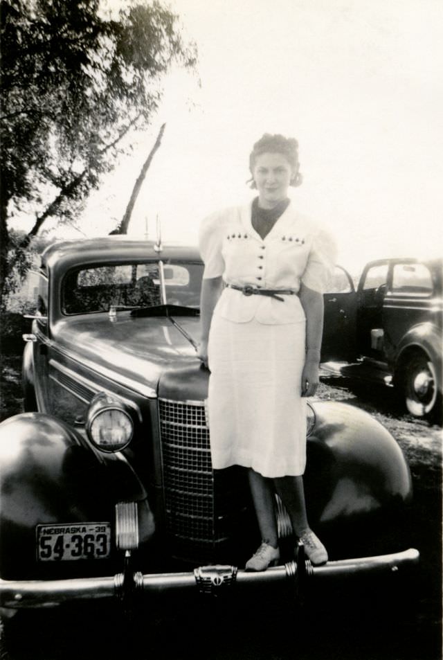 A stylish young lady in a white dress posing on the bumper of a 1937 Oldsmobile Eight Sedan in the countryside.