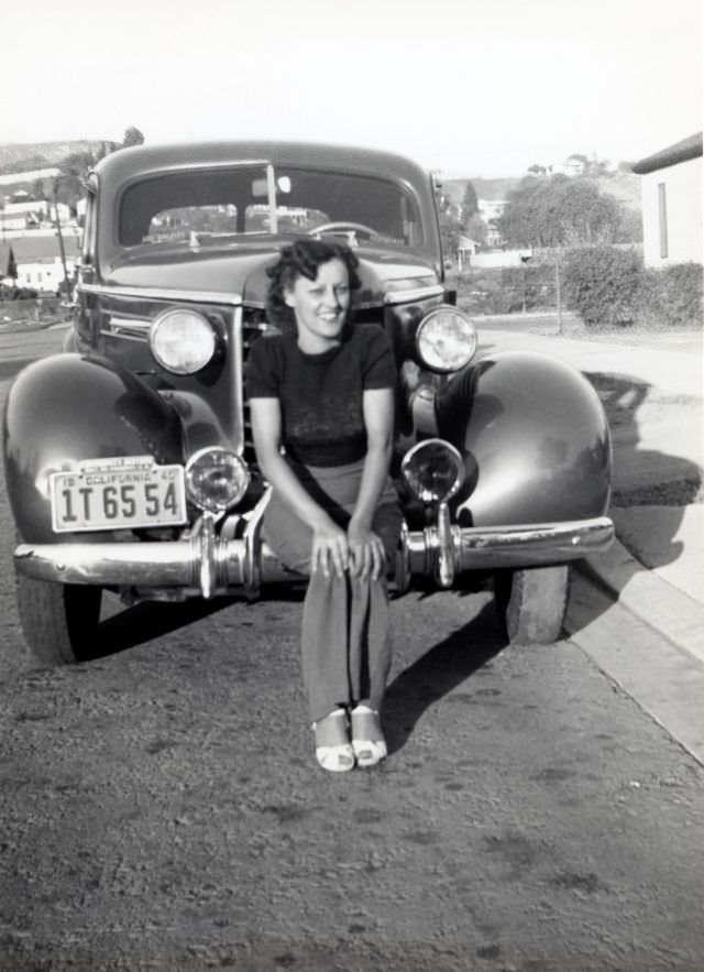 A cheerful young lady posing on the bumper of a 1937 Oldsmobile Six in a suburban street.