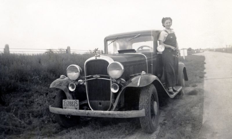 A young lady in bib overalls posing on the running board of a 1932 Oldsmobile Eight in the countryside.