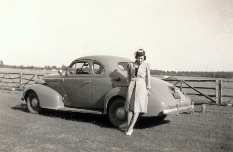 A young lady wearing a fashionable knee-length coat, extravagant hat, and white pumps bashfully posing with a 1937 Oldsmobile Business Coupe in the countryside, 1940