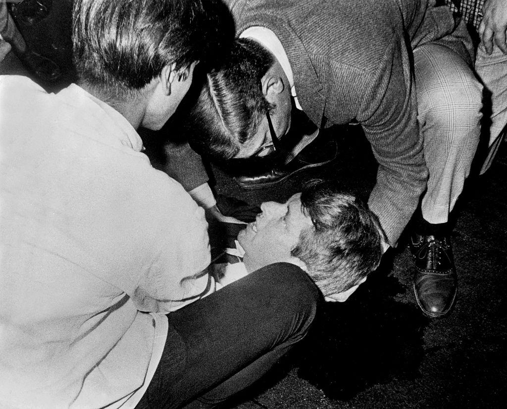 Robert F, Kennedy lies on the floor, his shirt is opened and he looks up at people assisting him with a pool of blood beneath.