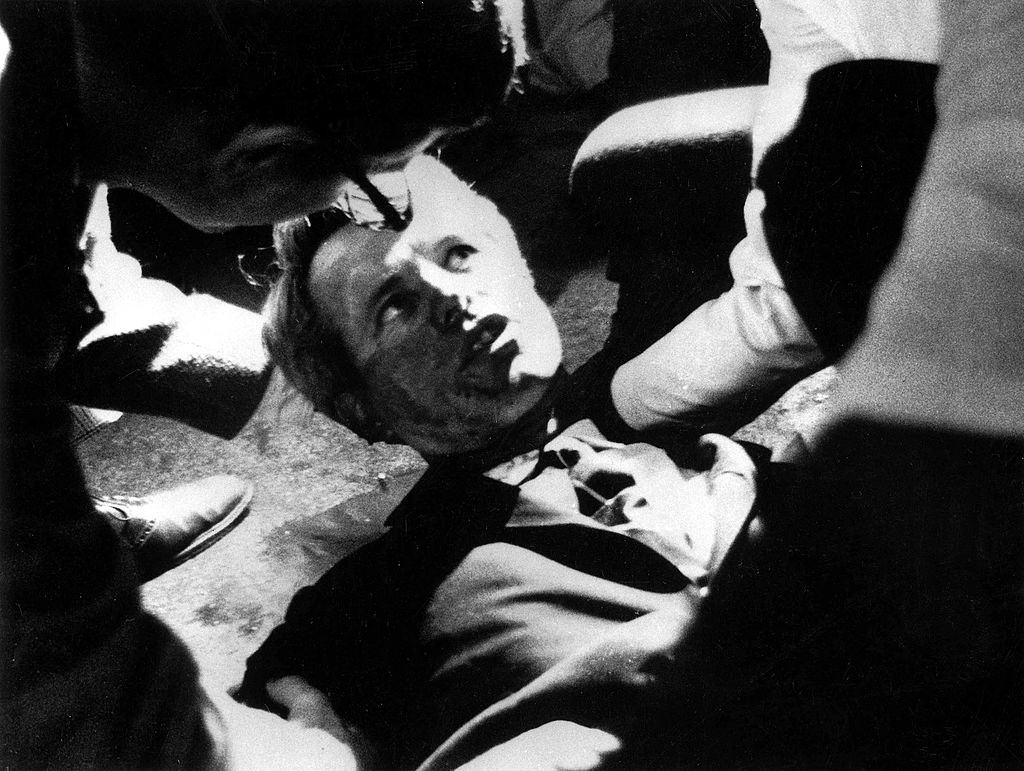 Robert F, Kennedy lies on the floor of the Ambassador hotel, his shirt is opened and he looks up at people assisting him.