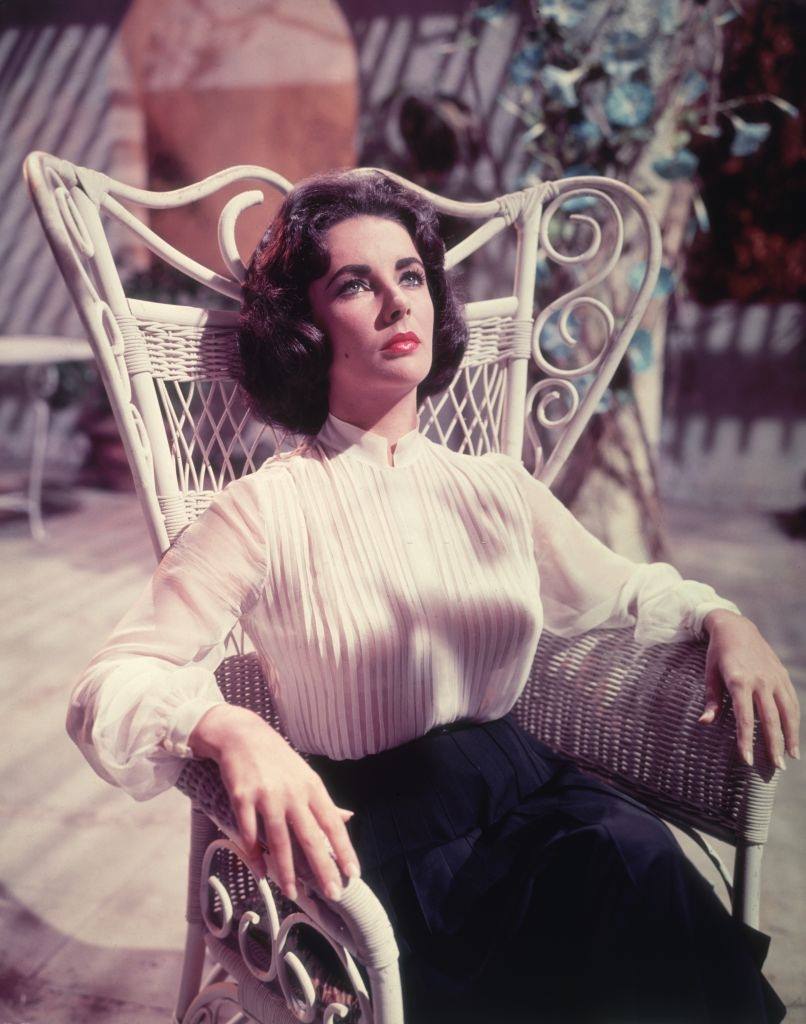 Elizabeth Taylor seated in a wicker patio chair in a still.