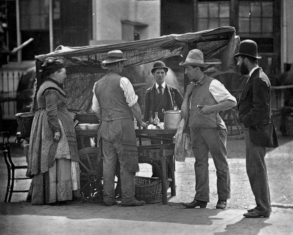 A shellfish stall owner sells oysters and whelks, 1877.