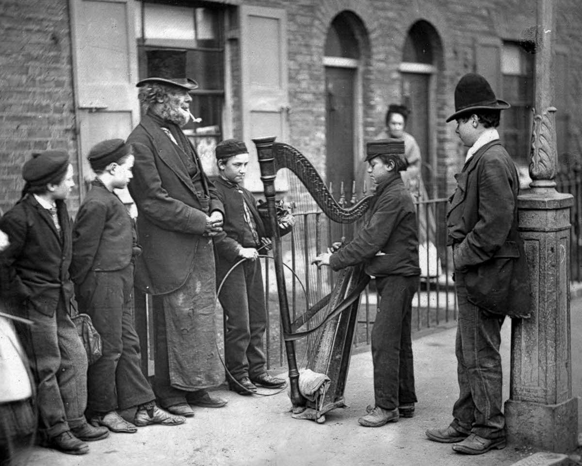 An Italian harpist entertains local children on the street, 1877.