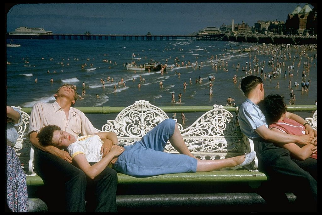 Young woman lying on white wrought iron bench w. head resting in lap of boyfriend in Blackpool beach, 1955.