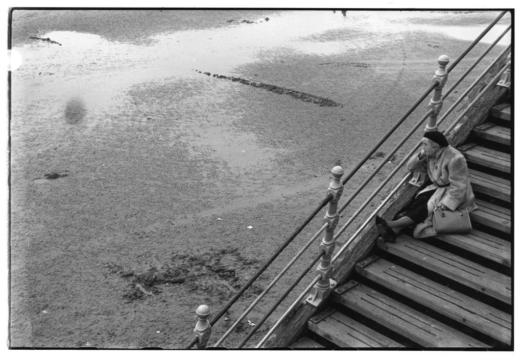 An elderly woman sits daydreaming on a flight of beachside steps, 1954.