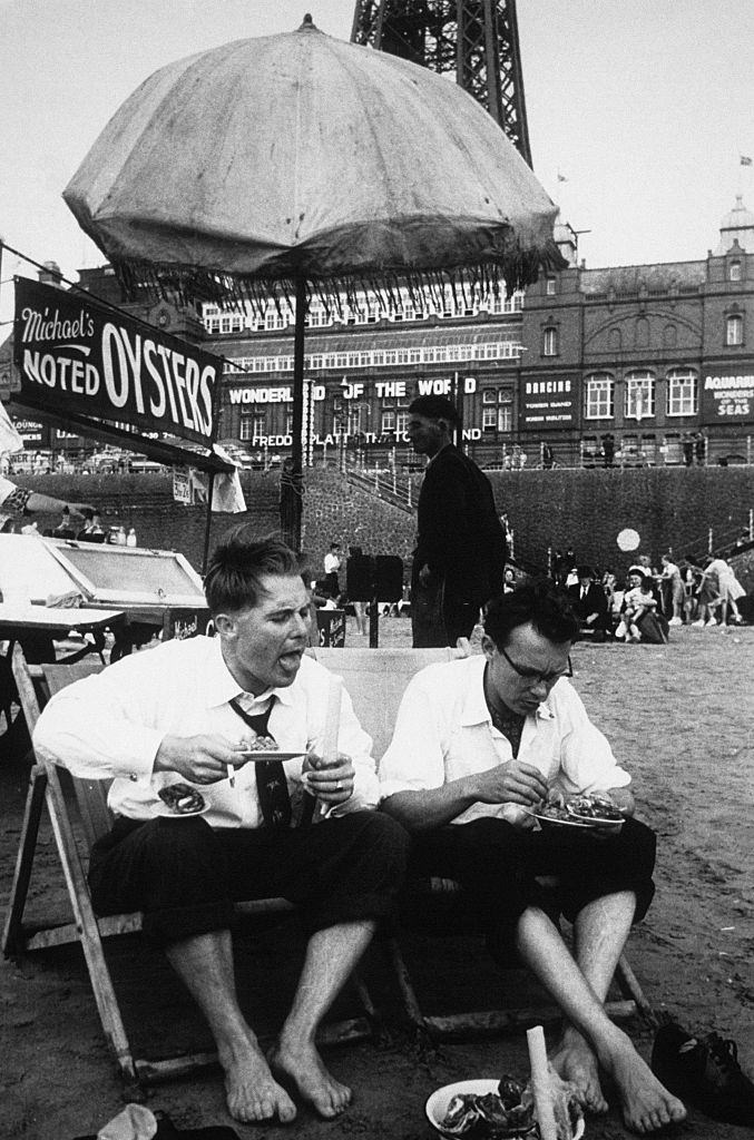 The comedians Eric Morecambe and Ernie Wise, enjoying some of the local delicacies on the beach at the foot of the Blackpool Tower, 1953.