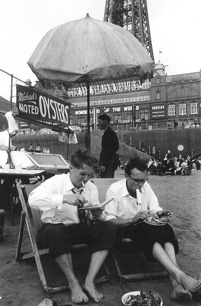 British comedy double act Eric Morecambe and Ernie Wise, in rolled up tousers, eating seafood on the beach at Blackpool, 1953.