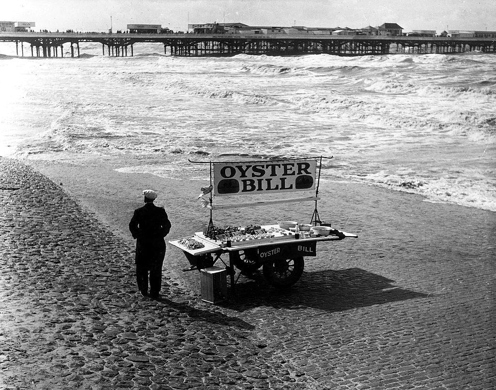 A man standing with a barrow displaying "Oyster Bill" on the sand at Blackpool, 1951.