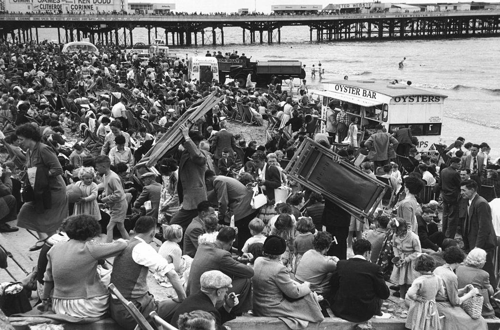 A crowd of people on the beach at Blackpool, some carrying deckchairs, July 1956.