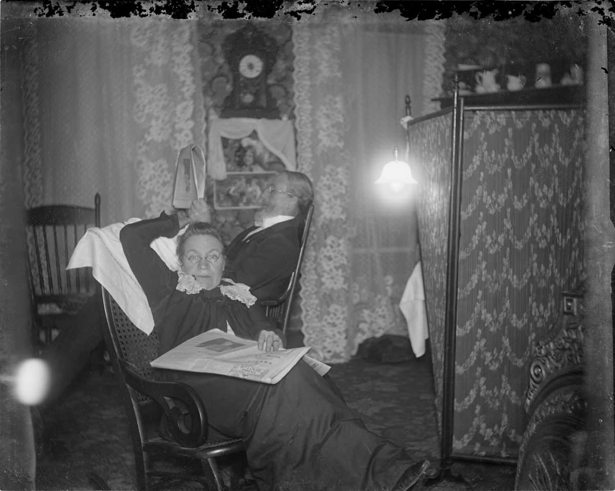 Woman sitting in rocking chair with newspaper on lap and man seated behind her – George Silas Duntley Photographs 1899-1918