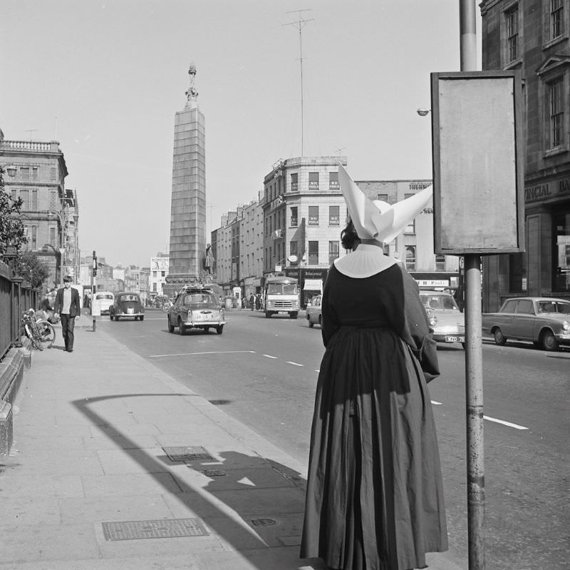 A nun with a most remarkable (and slightly dangerous-looking) veil/head dress waiting for a bus outside the Rotunda Hospital on Parnell Street, Dublin, 1964