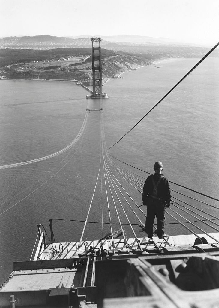 A man standing on the first cables during the construction of the Golden Gate Bridge with the Presidio and San Francisco, 1935.