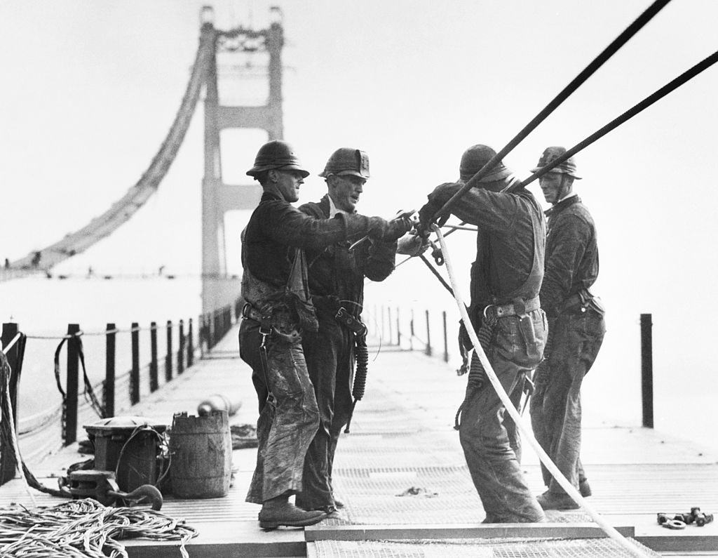 Workers are shown standing on a catwalk working on the cables of the Golden Gate Bridge spanning San Francisco Bay.