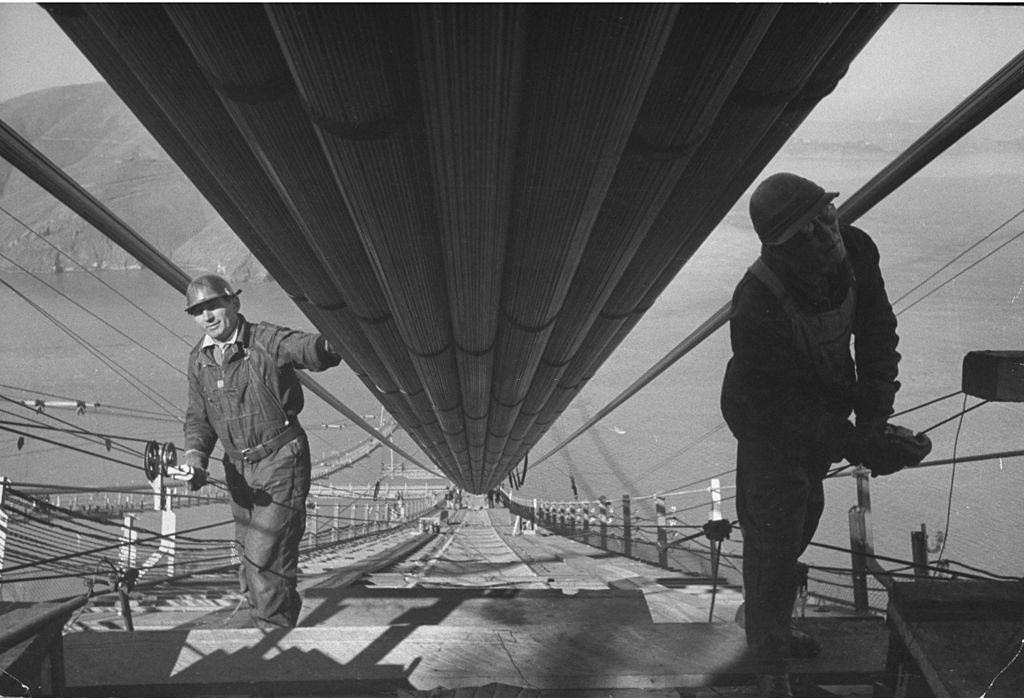 Two workmen adding the last two strands to one of the two enormous cables to the structure that supports a 6-lane highway, during construction of the Golden Gate Bridge.