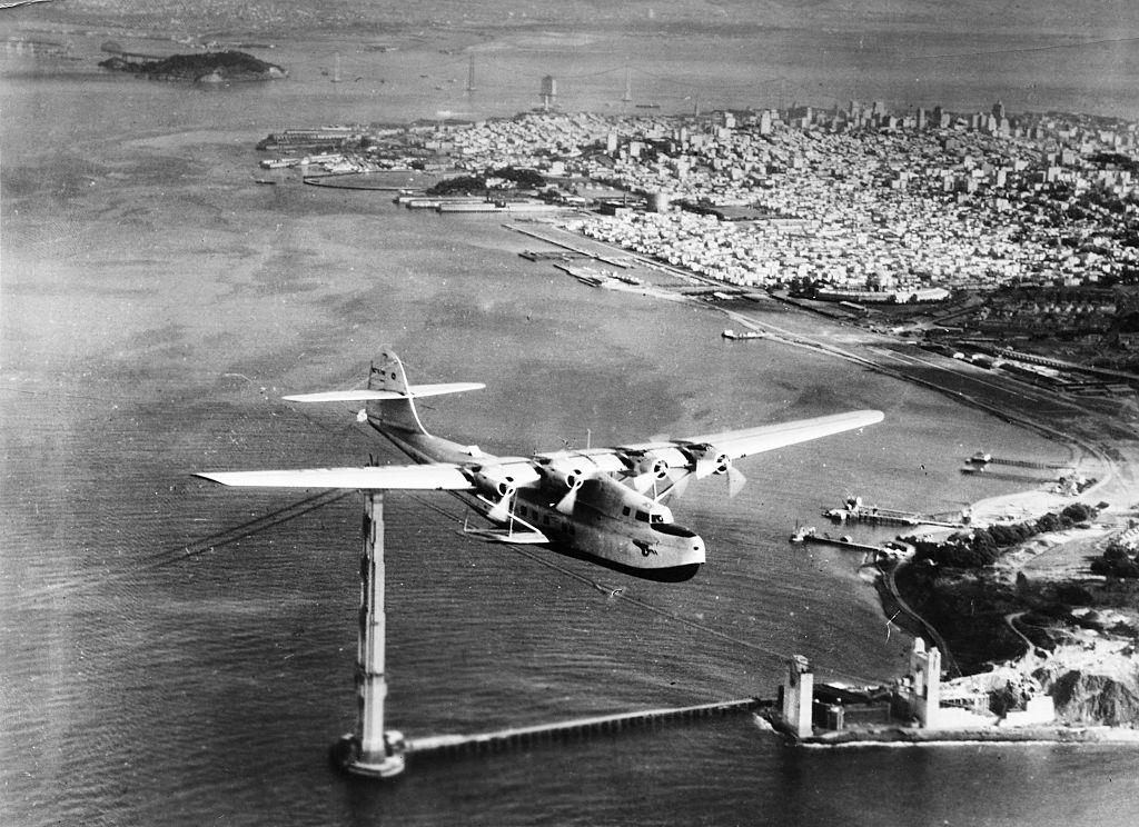 The China Clipper is shown flying over the incomplete Golden Gate Bridge.
