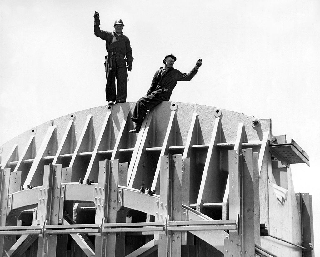 The Golden Gate Bridge under construction with two workers on the saddle atop the Marin Tower, 746 feet above the water, San Francisco, California, 1934.