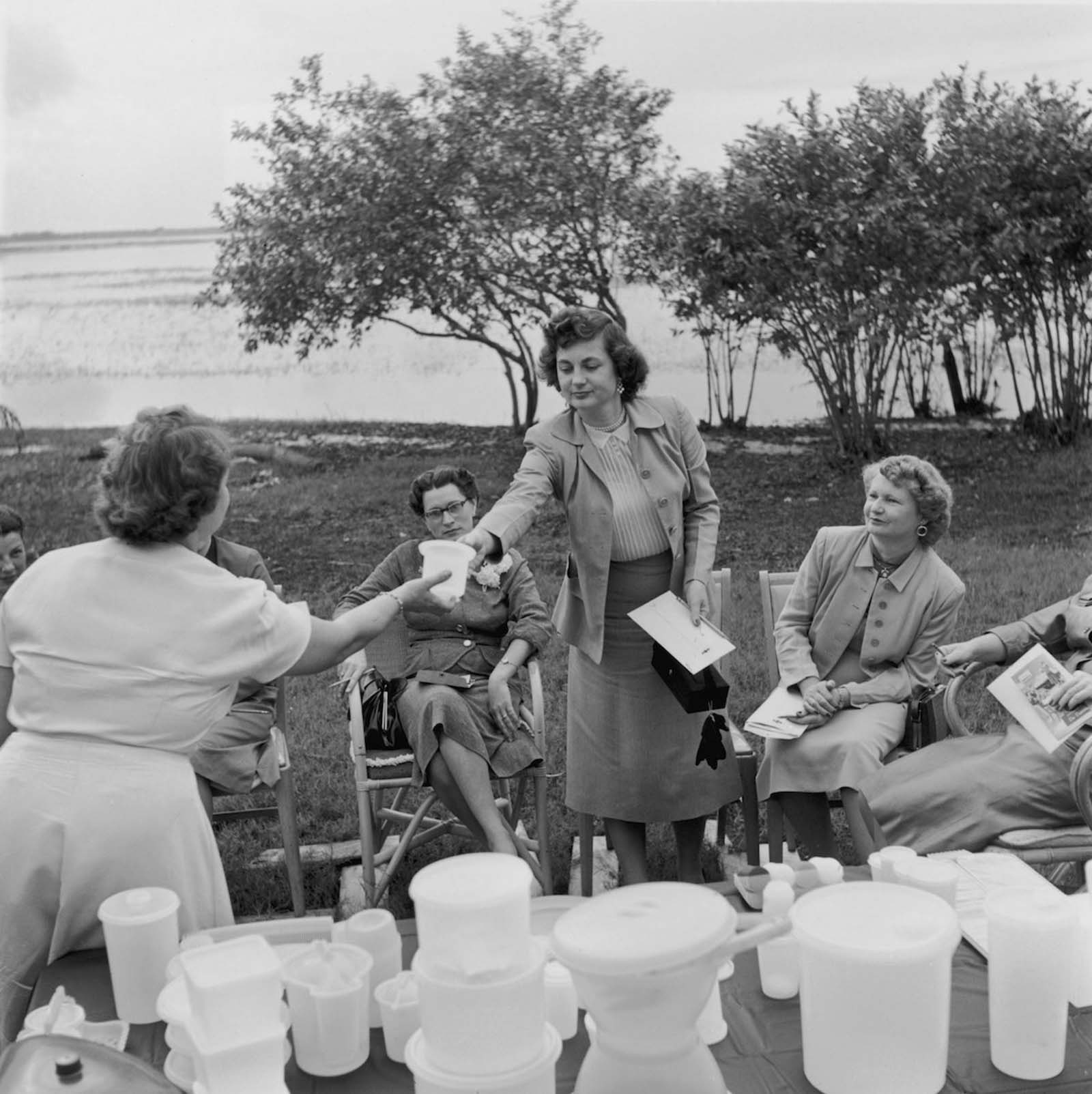 Passing round the product at an outdoor Tupperware. Hosts had a strict dress code – skirts and stockings were mandatory. 1955.