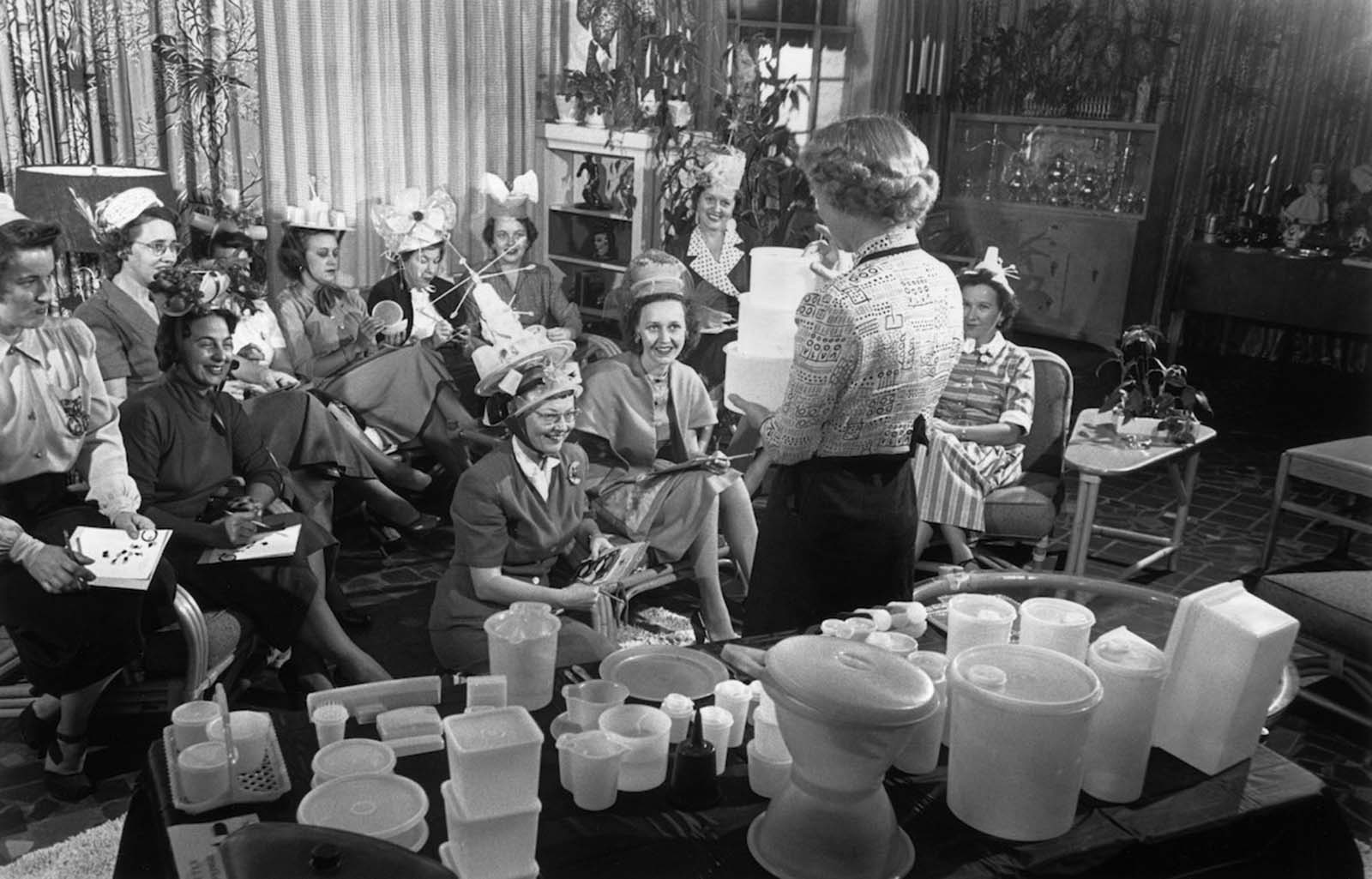 A Tupperware party in full swing. A table in the foreground displays a range of the company’s products. 1950s.