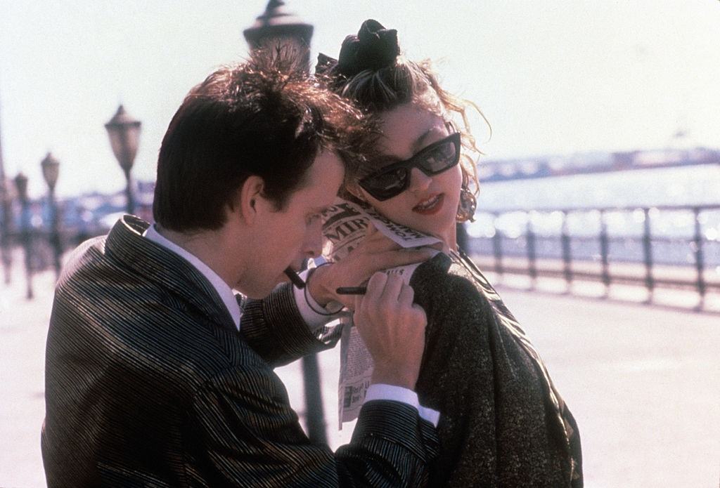 Canadian actor Robert Joy writing a note on a newspaper resting on Madonna, 1985.