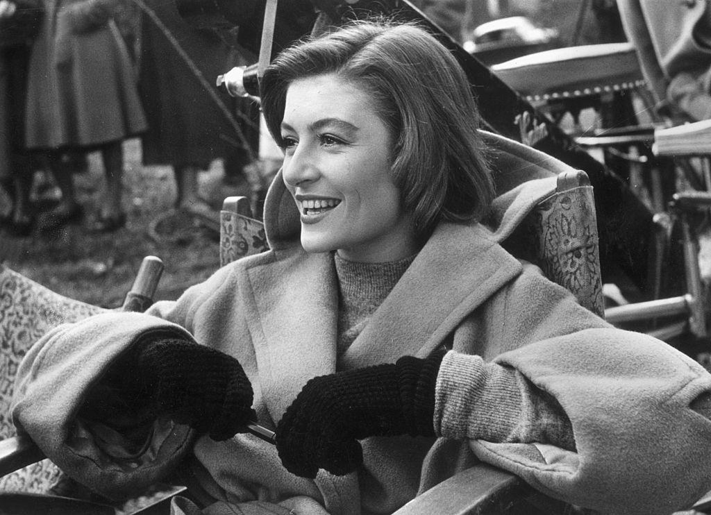 Anouk Aimee, wearing a camel-hair overcoat and gloves, laughs while sitting in a folding chair on a film set, 1955.