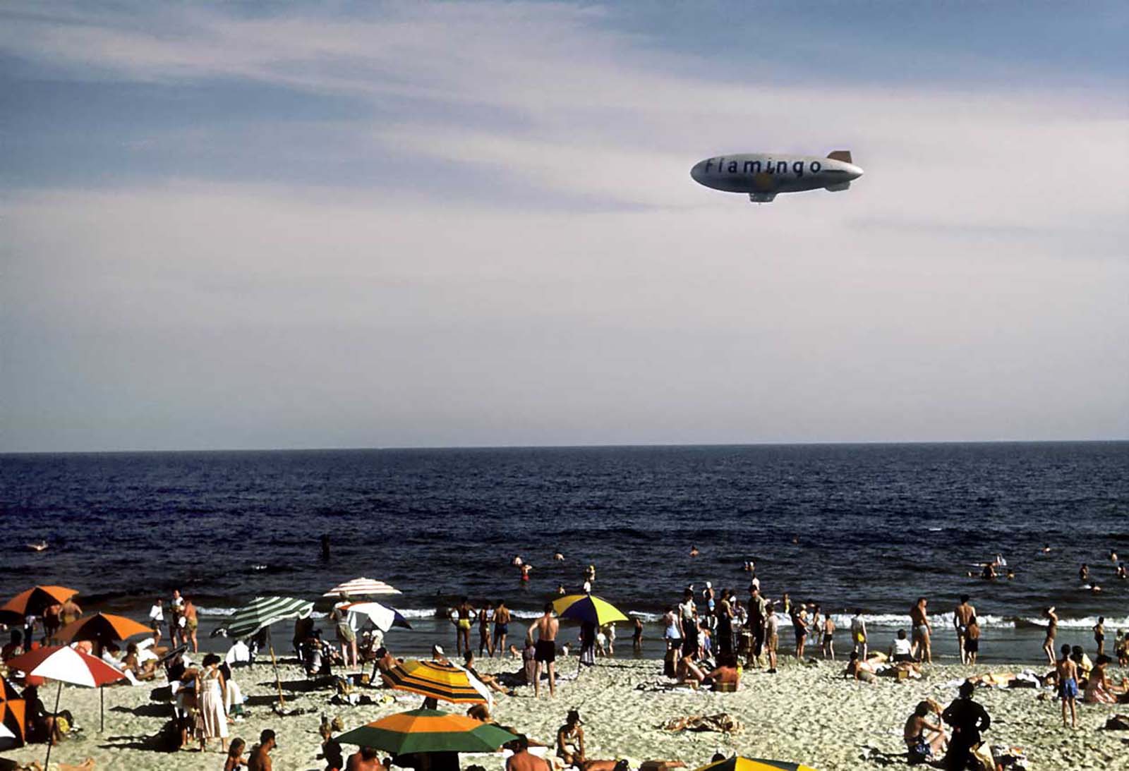 Sunbathers and swimmers on the beach while a blimp with the word Flamingo on the side glides above.