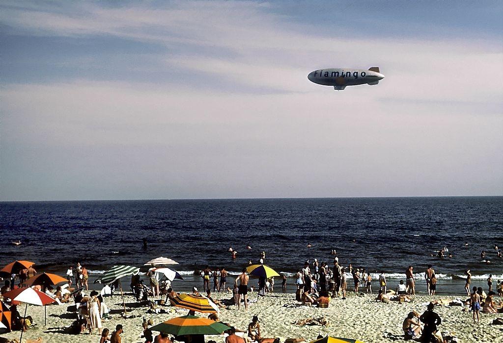 Sunbathers and swimmers frolic on Coney Island beach while a blimp with the word Flamingo on the side glides above, 1948.